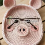 A crocheted pink pig face dish with round glasses placed over it, sitting on a wooden surface with small flowers in the background.