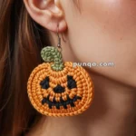 A close-up of a woman's ear wearing a crochet pumpkin earring with a smiling face design, featuring black eyes and mouth, with a green stem loop at the top.