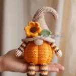 Hand holding a crocheted doll designed as a gnome with a pumpkin body. The doll has a pointed hat adorned with a yellow flower.