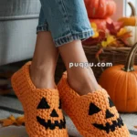 Person wearing orange crochet slippers with jack-o'-lantern faces, surrounded by autumn leaves and pumpkins.