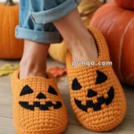 A person wearing orange crocheted slippers with black jack-o'-lantern faces on a wooden floor, surrounded by autumn leaves and pumpkins.