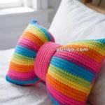 A colorful, bow-shaped pillow with horizontal rainbow stripes on a white bed.