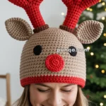 A person wearing a crocheted reindeer hat with red antlers and a red nose, smiling in front of a blurred Christmas tree.