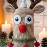 A crochet cover designed as a reindeer with antlers and a red nose, wrapped around a soap dispenser bottle, set against a festive background with candles and pinecones.