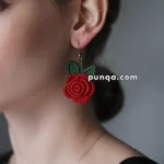 A person wearing a red rose-shaped earring with green leaves, made from beaded fabric, shown in profile against a plain background.