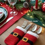 A festive table setting featuring two crocheted red cutlery holders resembling Santa's suit, complete with black belts and yellow buckles, placed beside a set table with plates, glasses, and red candles on a green and red tablecloth.