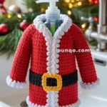 A soap dispenser dressed in a knitted Santa Claus outfit, complete with a red jacket, black belt, and white trim, sitting on a kitchen sink with festive decorations in the background.