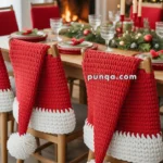 A festive dining room with chairs covered in red and white knitted Santa hat chair covers, a decorated Christmas tree, a fireplace with stockings, and a table set for a holiday meal with candles and greenery.