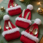 Red crocheted Santa hat napkin holders with cutlery inside, featuring white fluffy pom-poms and trim, displayed on a green tablecloth with Christmas decorations such as pine branches, bells, and string lights.