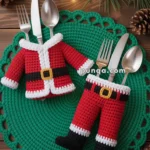 Forks and spoons are placed in crocheted Santa suit holders, featuring a red jacket and pants with white trim, lying on a green crocheted mat with pine branches and lights in the background.