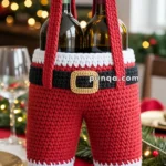 Two wine bottles are held upright inside a crocheted holder designed as Santa Claus's pants and boots with suspenders, on a festive dining table with holiday decorations.