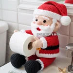 A crocheted Santa Claus figure holding a roll of toilet paper, sitting on the edge of a bathroom sink with a small decorative star and a sprig of pine nearby.
