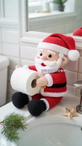 A crocheted Santa Claus figure holding a roll of toilet paper, sitting on the edge of a bathroom sink with a small decorative star and a sprig of pine nearby.