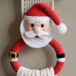 A crocheted Santa Claus towel holder hanging from a metal rod, with Santa's face and hat crocheted at the top above a red ring adorned with a faux belt buckle. A white towel is draped through the ring.