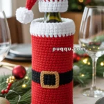 A wine bottle covered with a crocheted Santa Claus outfit, featuring a red suit with a black belt and a Santa hat, placed on a festive table setting with holiday decorations.