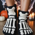 Person wearing knitted black slippers with white bone designs, resembling a skeleton foot pattern, on a wooden floor with a Halloween-themed background including pumpkins and candles.