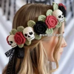 A woman with blonde hair wears a crocheted headband featuring a pattern of skulls, red and black flowers, and green leaves, with a lace ribbon tied at the back.