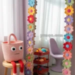 A room featuring a full-length mirror adorned with colorful crocheted flowers. Nearby, a pink crocheted basket with googly eyes and red legs sits on a wooden stool. A purple chair and a cart with various colored yarn balls are visible in the background.
