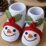 A pair of crocheted baby booties shaped like smiling snowmen, featuring red hats and green leaf accents, displayed on a wooden surface with festive decorations in the background.