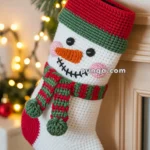 A crocheted Christmas stocking featuring a snowman design with a red, green, and white color scheme hanging from a decorated mantle with pine branches and Christmas lights.