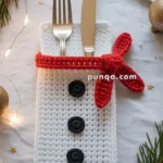 A fork and knife are placed in a crochet white pouch resembling a snowman, with black button details and a red scarf. The setting includes festive decorations like pine branches, gold baubles, and twinkling lights on a white tablecloth.