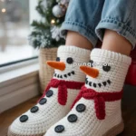 A pair of feet wearing crocheted snowman-themed slippers with orange carrot-like noses, black button eyes, and red scarves, beside a decorated Christmas tree.