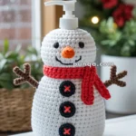 A soap dispenser disguised as a snowman with a crochet cover, featuring a carrot nose, red scarf, and black button details, sitting on a bathroom counter with a festive background.