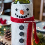 A wine bottle dressed in a crocheted snowman outfit, complete with button eyes, a carrot-shaped nose, and a red scarf. The bottle has a crocheted black hat with green leaves and red berries. The setting includes pinecones and holiday lights.
