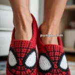 Feet wearing red crochet slippers designed to resemble a superhero mask with large white eyes.
