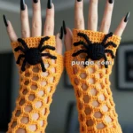 Two hands wearing orange crocheted fingerless gloves with black spider designs, displayed against a blurred indoor background.