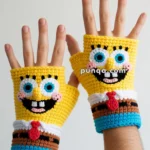 Two hands wearing crocheted fingerless gloves designed with a cartoon character face, featuring large blue and black eyes, a wide smile, and a red tie, against a plain background.