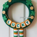 A green wreath decorated with crocheted clovers, gold coins, and a top hat, featuring the word 'LUCKY' and dangling striped legs with black shoes.