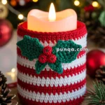 A candle wrapped in a red and white crochet cover adorned with green holly leaves and red berries, surrounded by festive decorations including pinecones, pine branches, and string lights.