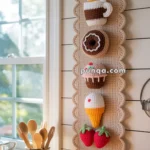 A crocheted wall hanging in a kitchen features designs of a steaming mug, donut, cupcake, ice cream cone, and strawberries on a beige scalloped cloth background.