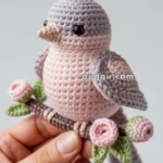 A hand holding a crocheted bird with grey and pink yarn, perched on a crocheted branch with pink flowers and green leaves.