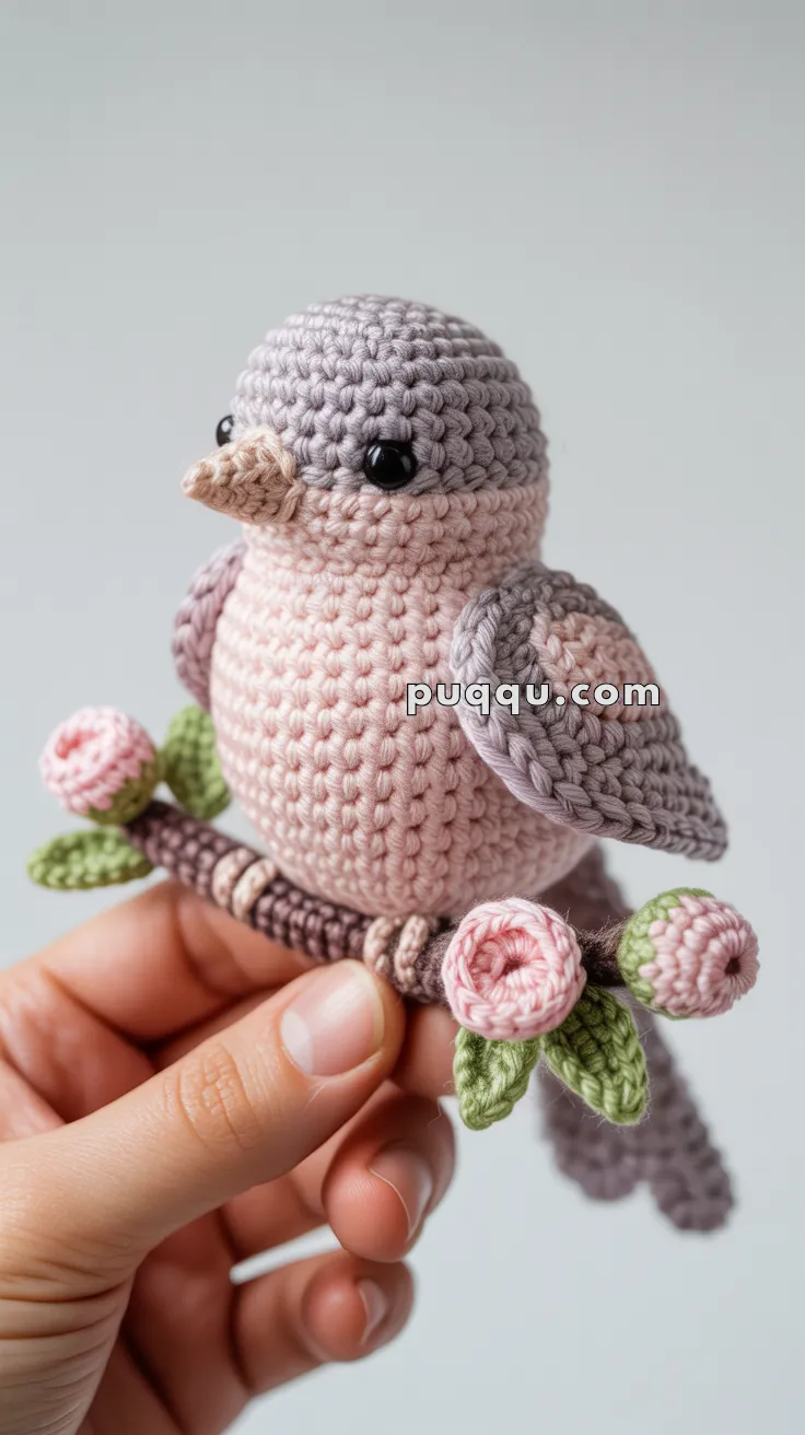 A hand holding a crocheted bird with grey and pink yarn, perched on a crocheted branch with pink flowers and green leaves.
