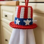 A crocheted kitchen towel holder shaped like a red, white, and blue top hat hanging from a cabinet knob, with a white towel draped underneath.