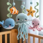 A crochet baby mobile hangs above a crib, featuring cute sea creatures like a whale, clownfish, jellyfish, starfish, and octopus in pastel colors.