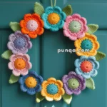A colorful crocheted floral wreath hanging on a teal door, featuring yarn flowers in shades of orange, pink, purple, blue, and green leaves.