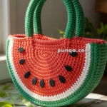A knitted watermelon-themed handbag with green handles, showcasing a red, white, and green pattern with black seed accents, placed on a rustic wooden surface near a window with natural light.