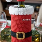 A wine bottle dressed in a crocheted Santa Claus costume with a red and white hat, red suit, black belt, and gold buckle, placed on a festive dining table.