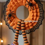 A crocheted Halloween wreath with an orange knit design, adorned with black spider decorations. Hanging from the wreath are striped orange and black legs ending in black shoes with purple bows, resembling a witch's legs. The background is a wooden door or window frame.